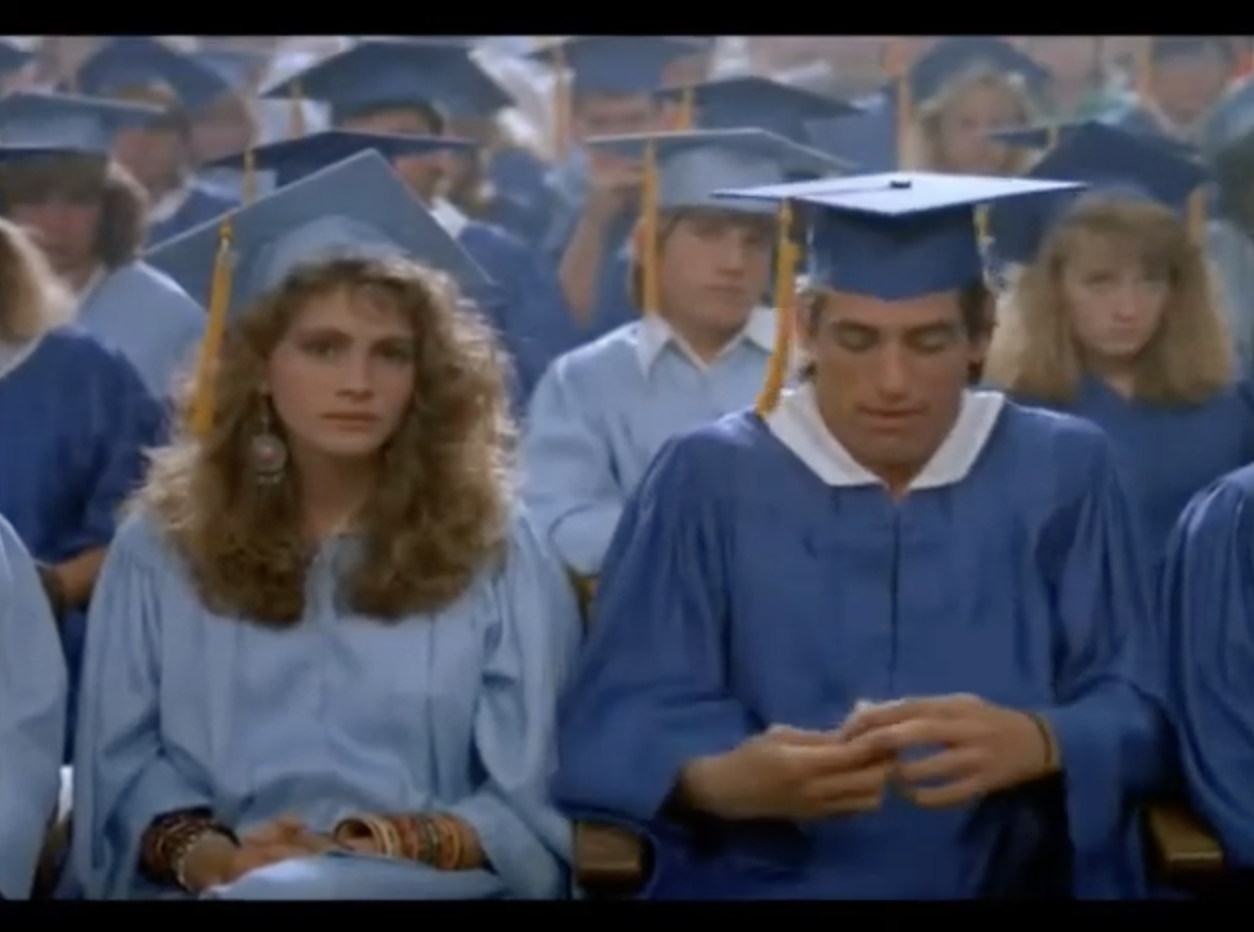 A young Julia Roberts sits in the front row of an audience, wearing a blue graduation gown and hat