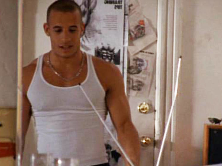 A young Vin Diesel stands in a cluttered apartment, wearing a tight white tank top