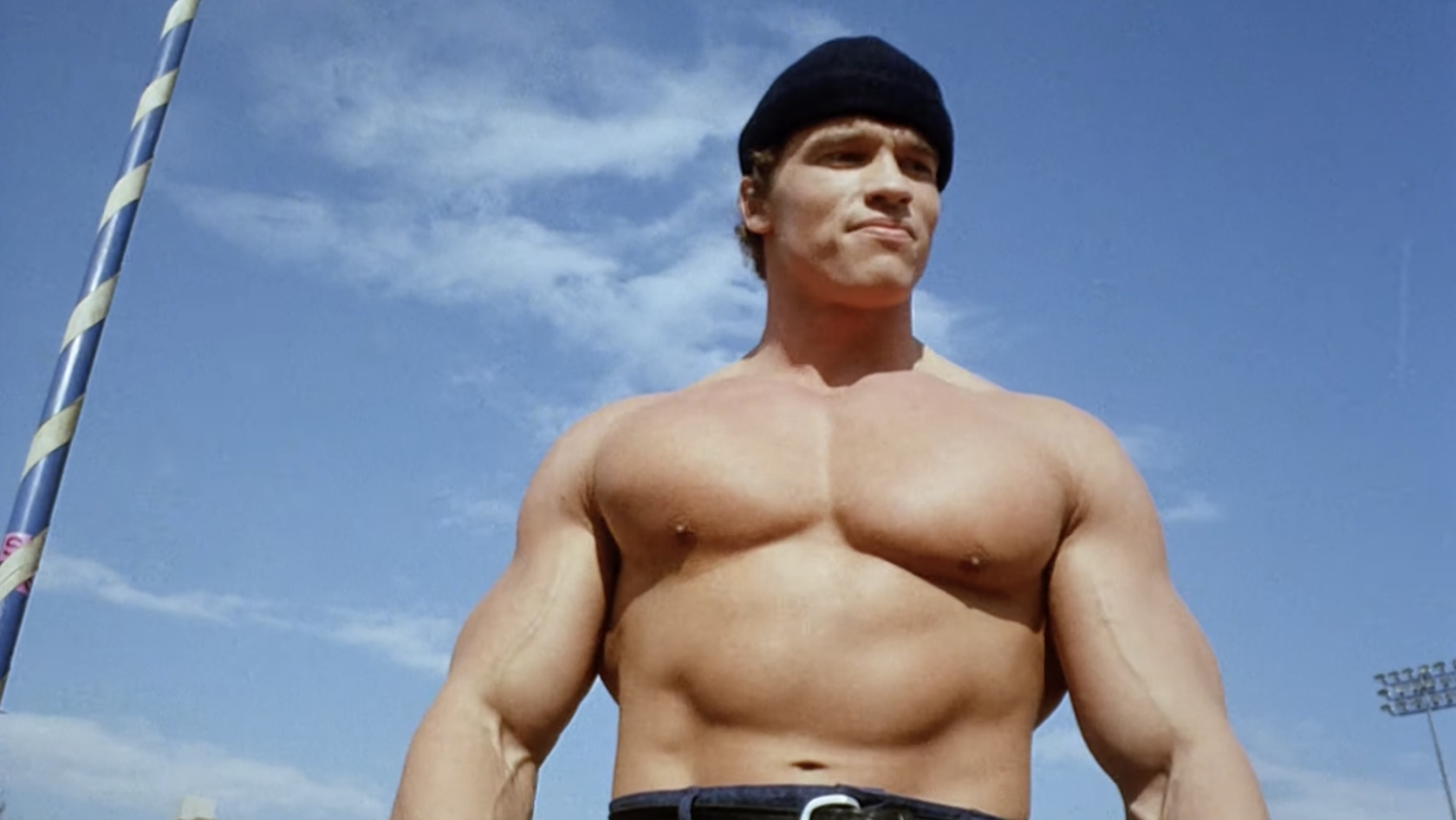 A young, shirtless, and very buff Arnold Schwarzenegger stands outdoors on a sport field wearing a beanie
