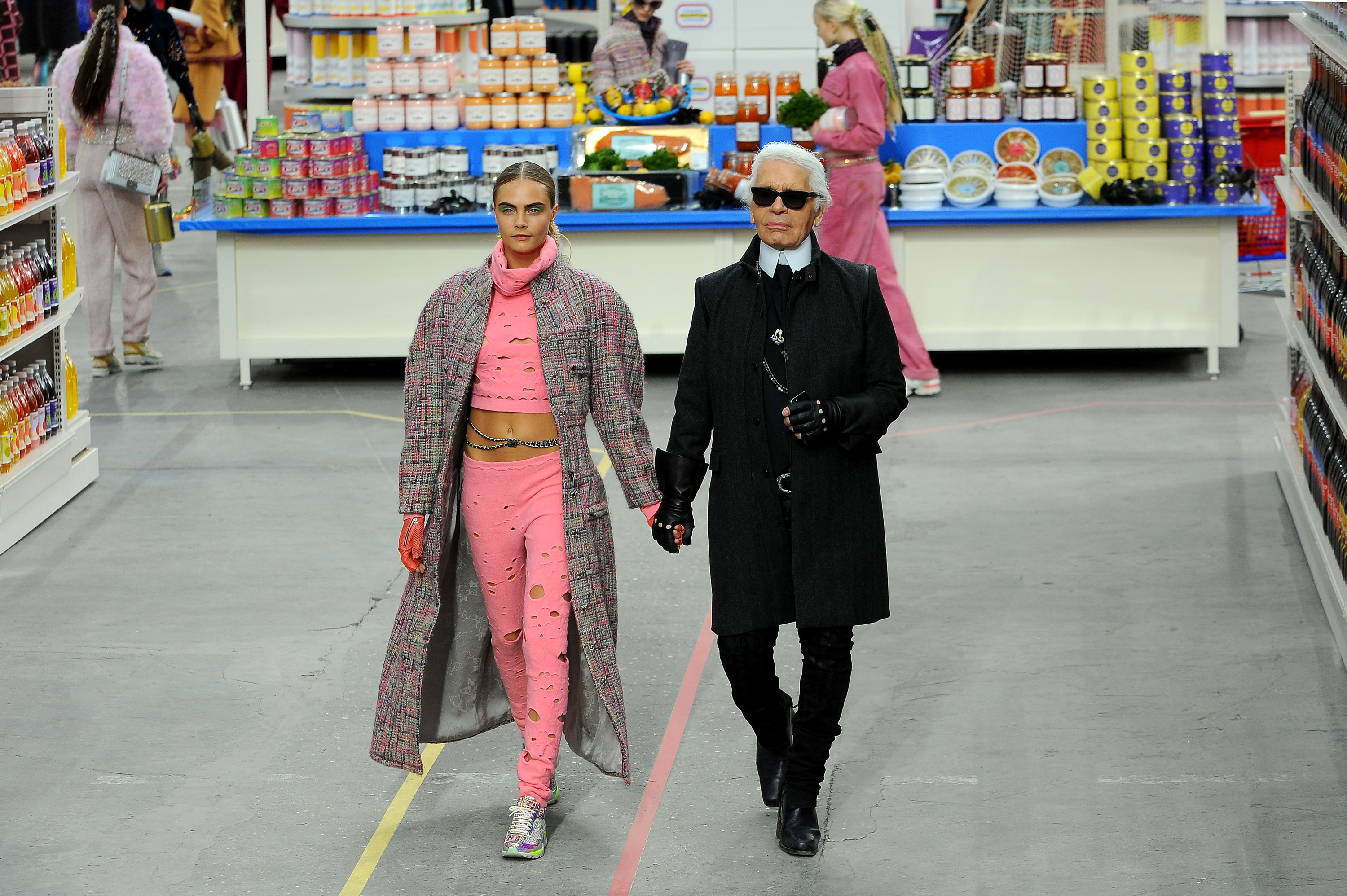 Karl walking down a runway designed to look like a grocery store aisle while holding hands with model Cara Delevingne