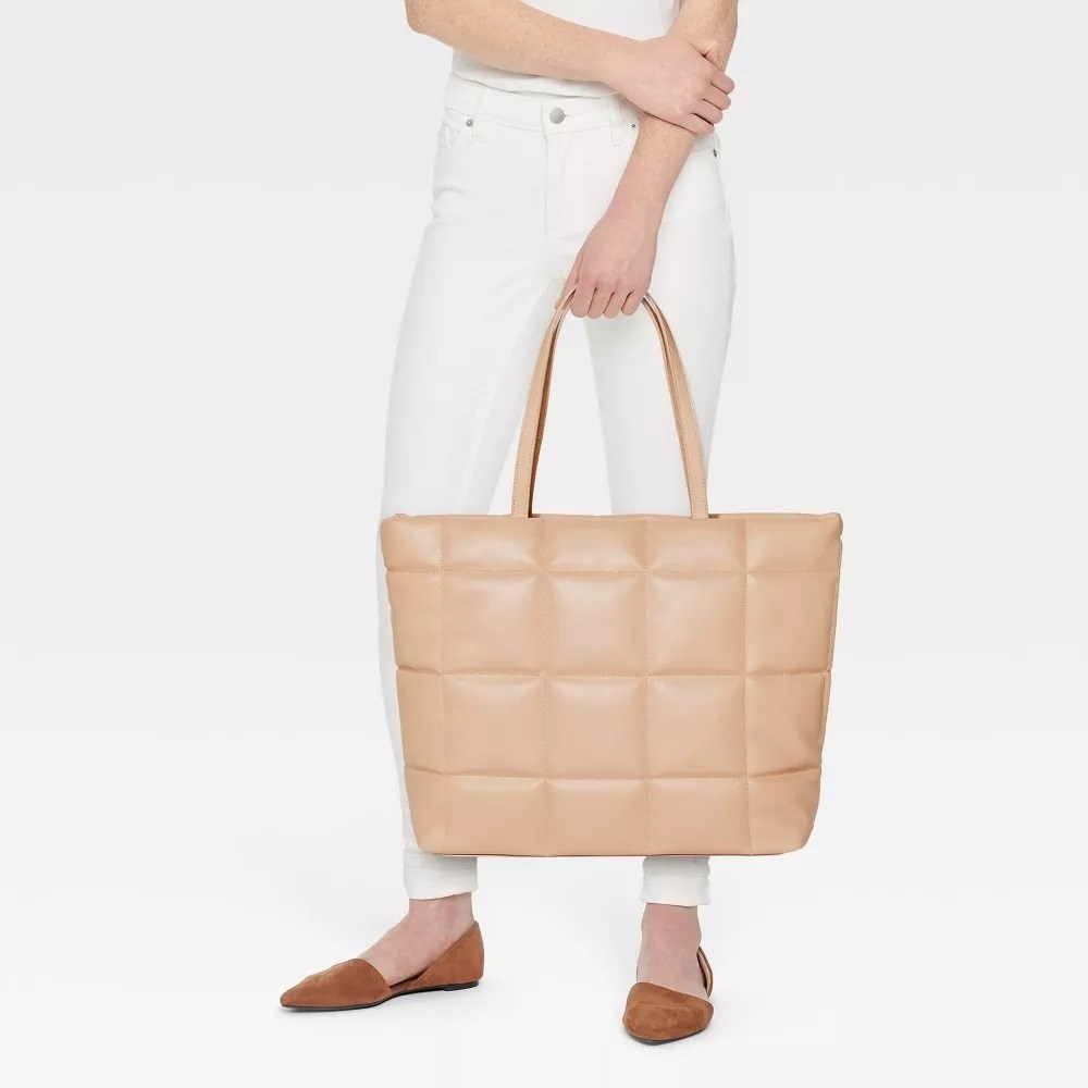 A model wearing white pants and holding the quilted tote in beige