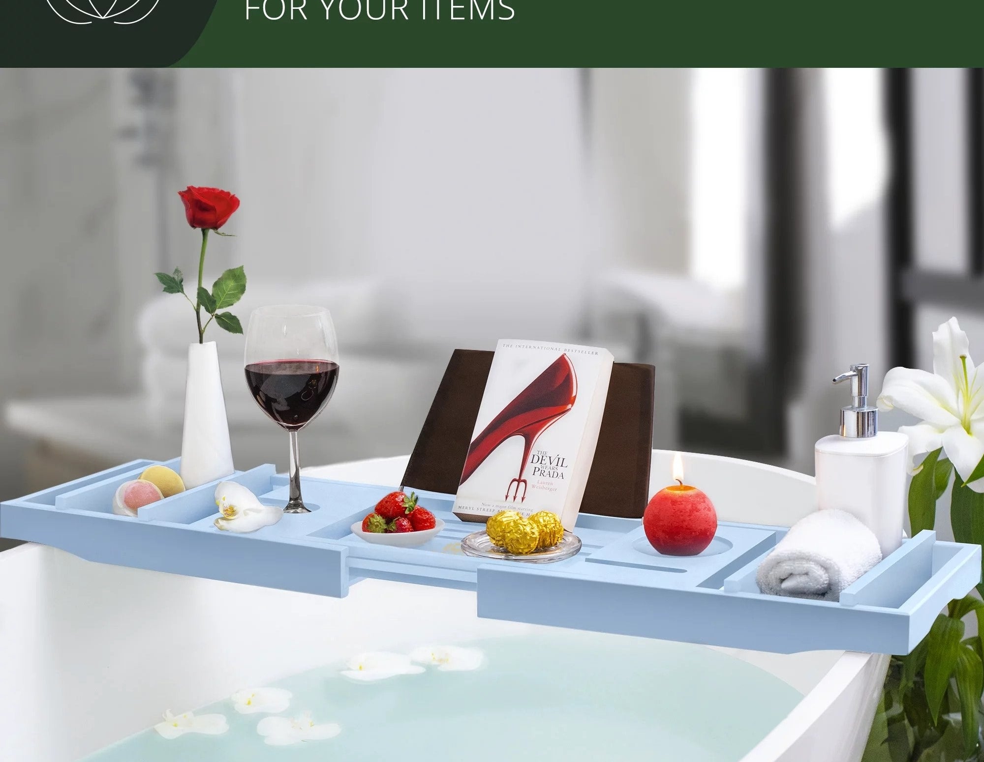 Baby blue bath tray holding multiple items including flower vase, glass of red wine, book, candle, soap dispenser and white towel, sitting over white tub