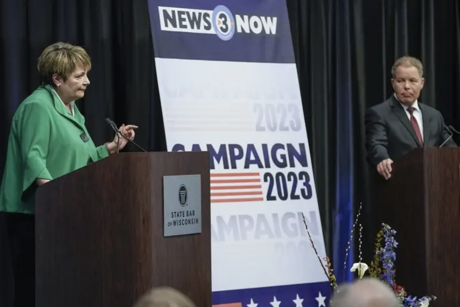 Dan Kelly and Janet Protasiewicz debate each other onstage in between a giant sign that reads &quot;campaign 2023&quot;