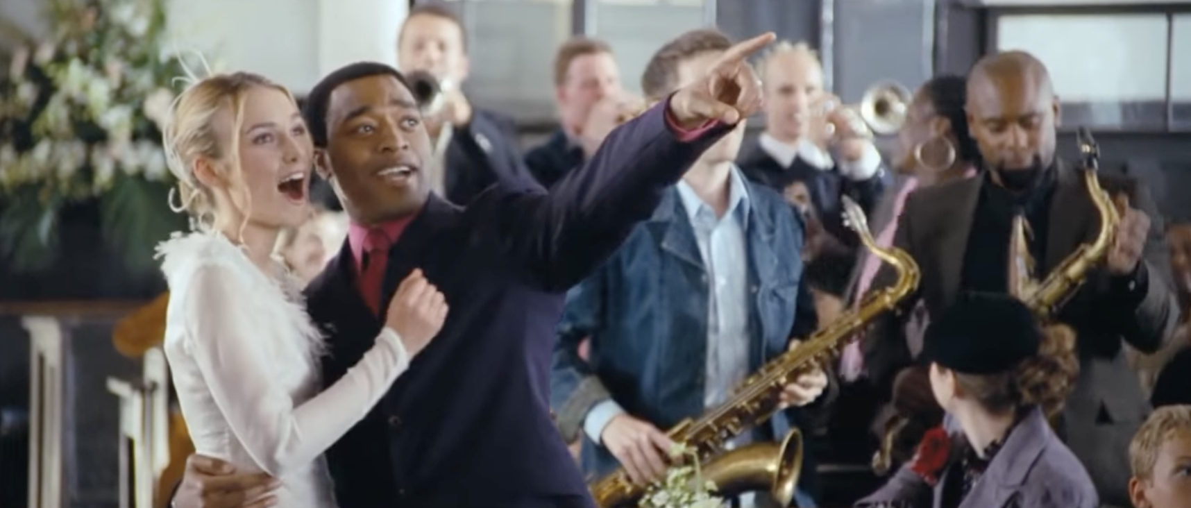 Juliet and Peter, in a white dress and a suit, stand in the aisle of a church pointing and exclaiming - two men with saxophones are seen behind them