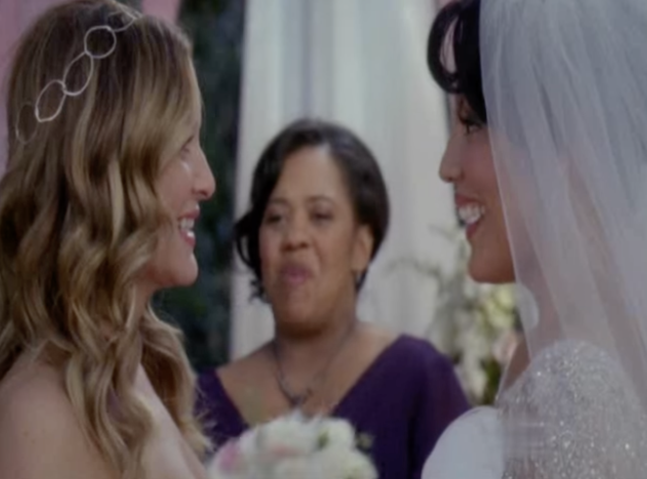 Callie, wearing a veil, and Arizona, holding flowers, stand face to face smiling, as Bailey is seen officiating between them in the background