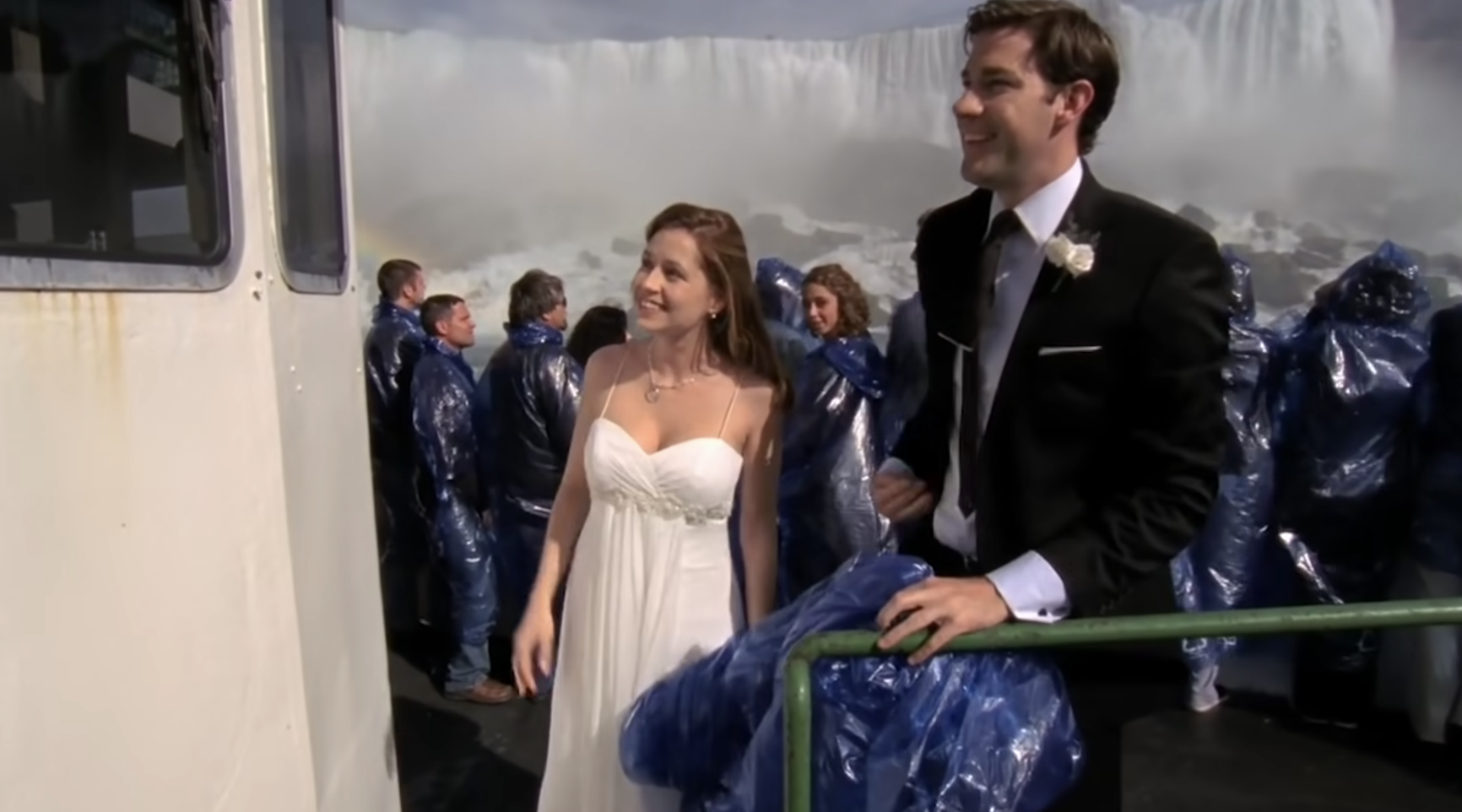 Jim and Pam stand on the deck of a boat holding blue plastic ponchos, a raging waterfall is seen behind them