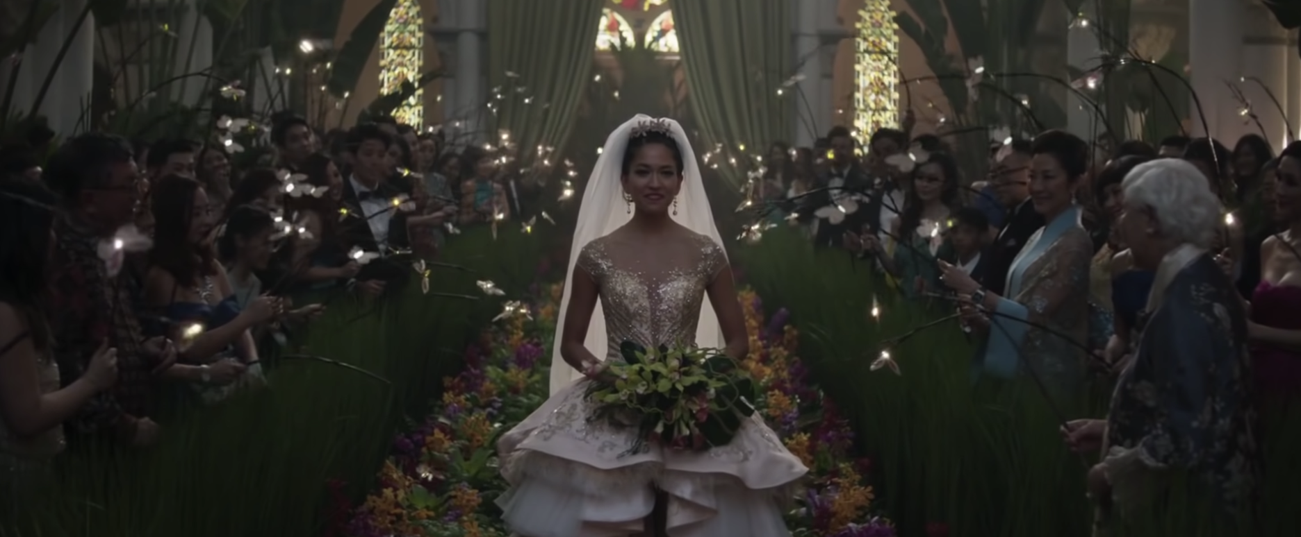 A bride walks down the aisle of a church holding a large bouquet of flowers, guests are holding glowing orbs on sticks