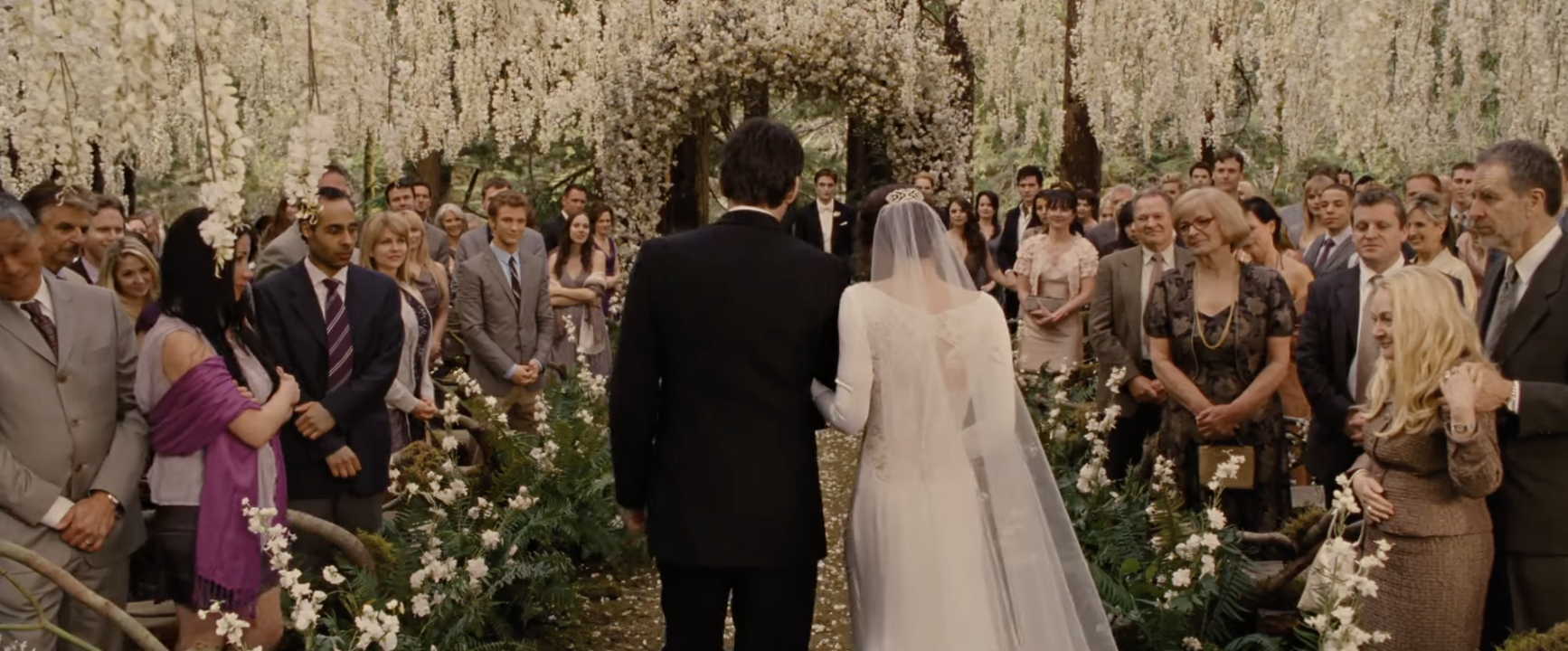 A bride and her dad are seen from behind, walking down a wedding aisle lined with flowers and greenery, white wisteria flowers hang above them