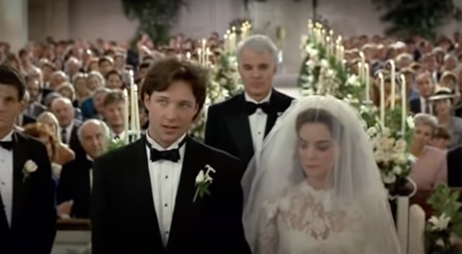 A shot of a bride and groom standing at the altar with the church at their backs, she has a veil over her face and he&#x27;s in a tux. Steve Martin, also in a tux, is standing behind them