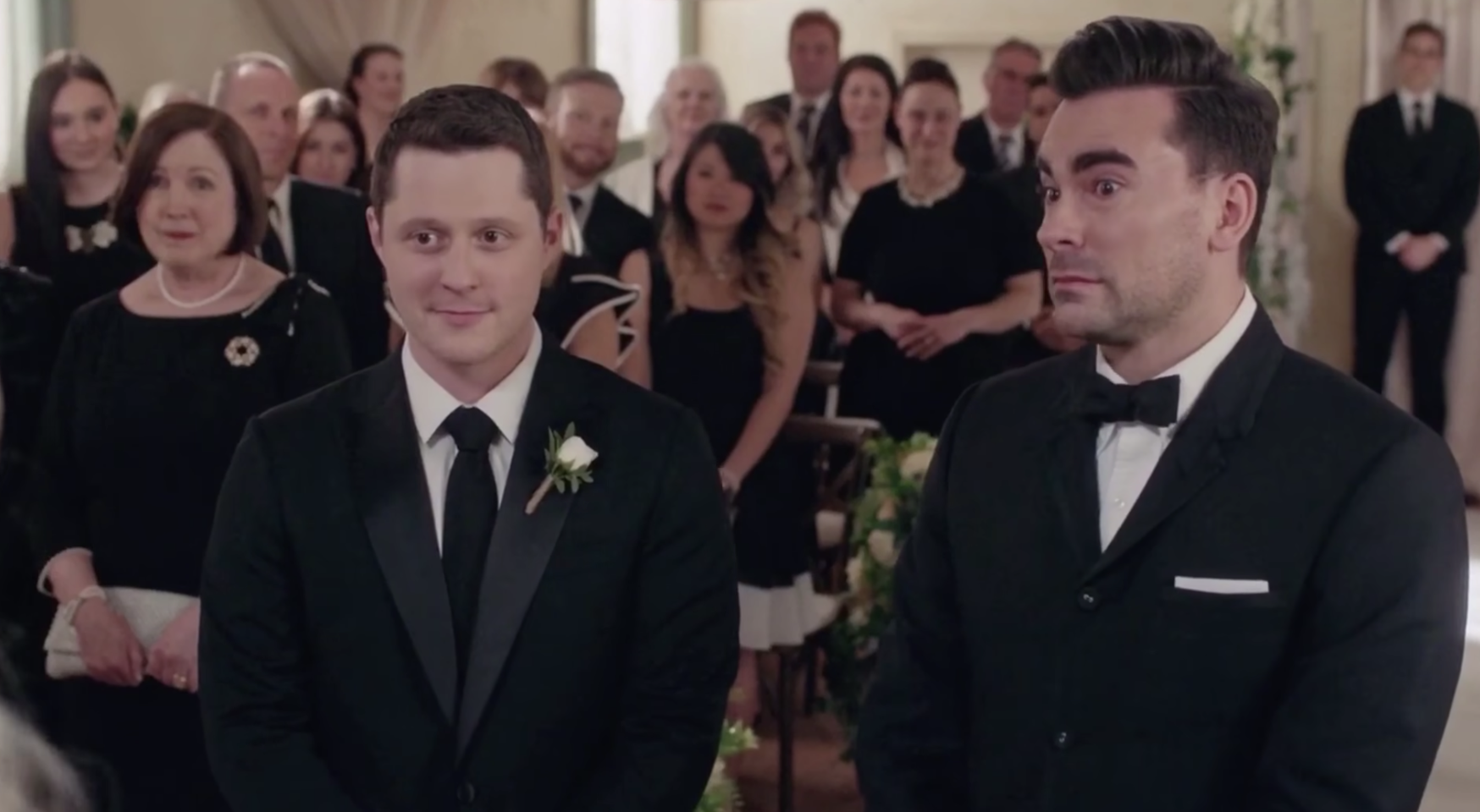 Patrick and David, both in black suits, stand at the altar with the church at their backs - the congregation is standing and all dressed in black