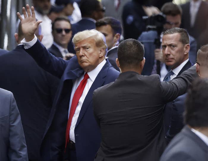 Donald Trump surrounded by men in suits has a solemn expression and waves to the camera in the middle distance