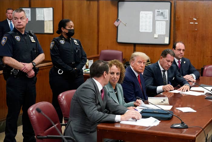 Donald Trump, flanked by two people on each side of him in suits, sits at a table and looks toward the camera with two police officers, one masked, standing behind him