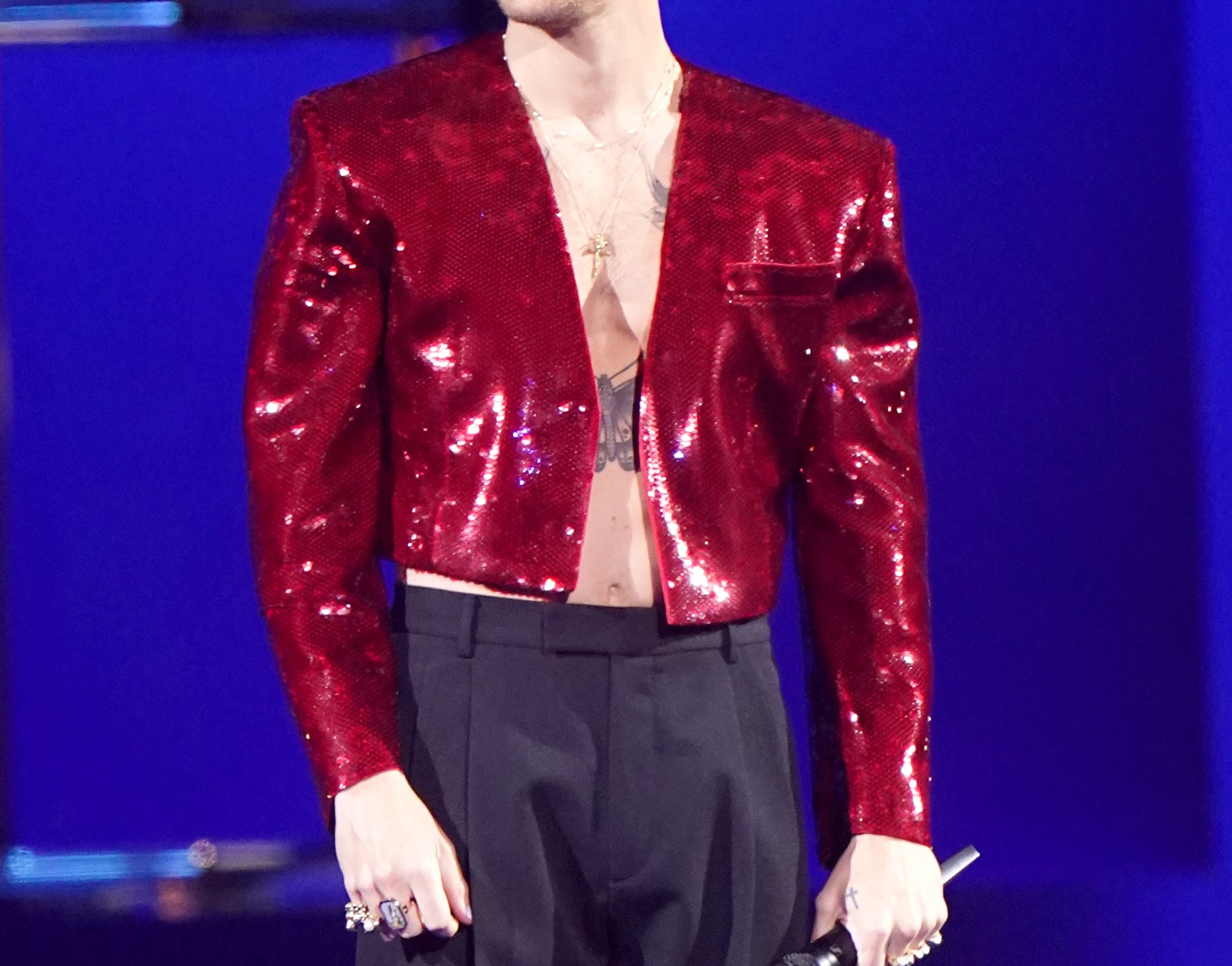 A close-up of Harry smiling in a shiny cropped jacket and bare-chested