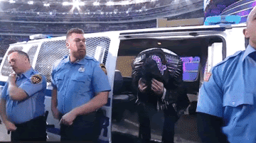 A masked Dominik Mysterio emerges from a police van