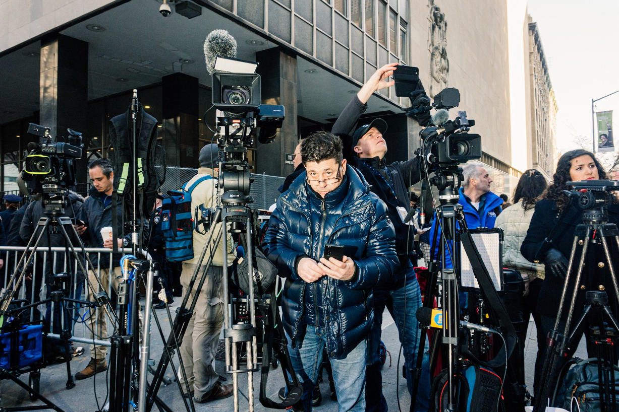 a member of a news crew looks at the camera as other crew members arrange cameras and lighting