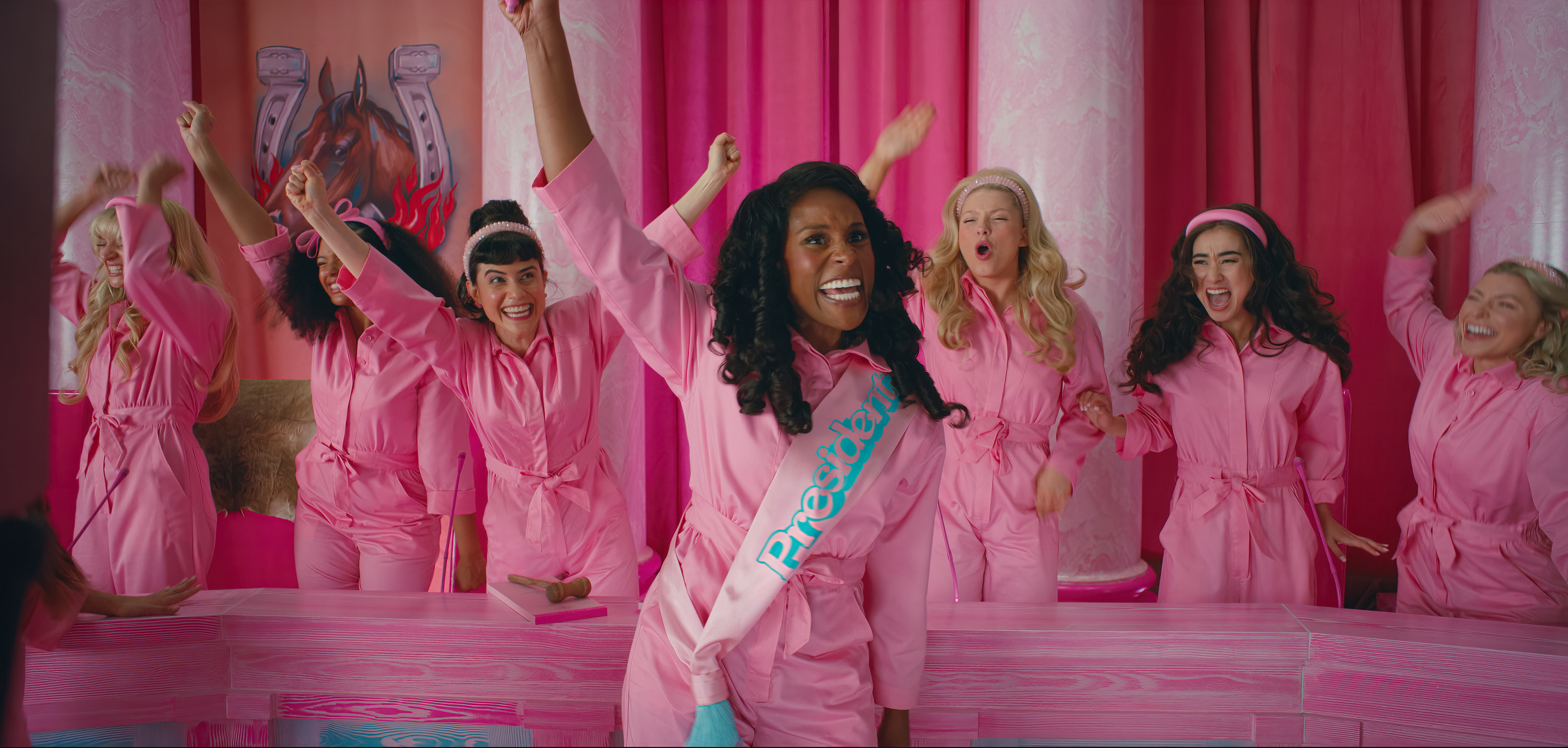 Issa Rae as Barbie riling up a crowd of people all wearing matching Barbie pink jumpsuits