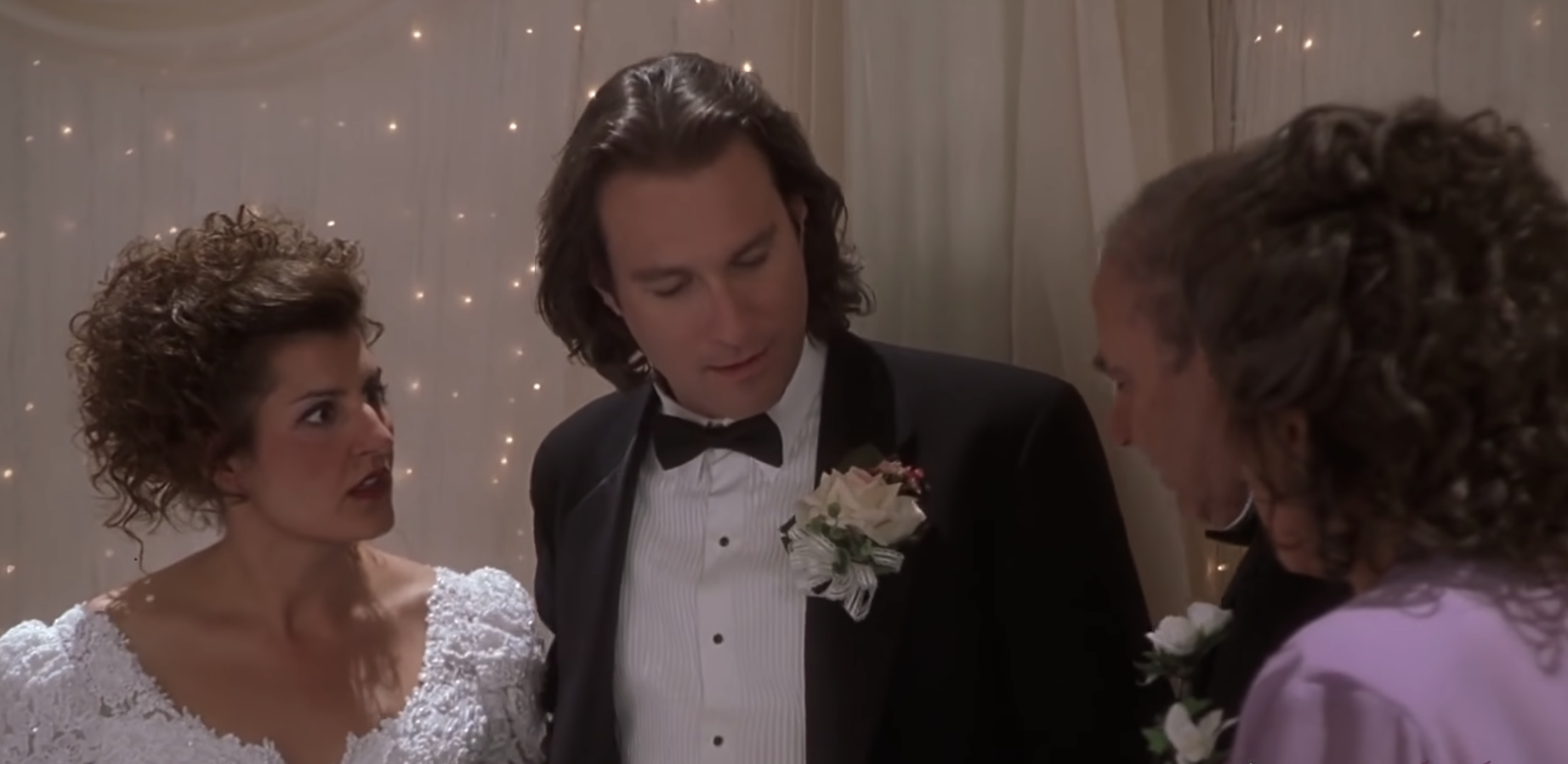 Toula, in a wedding dress, and Ian, in a black tux, stand in front of a wall of fairy lights, looking shocked