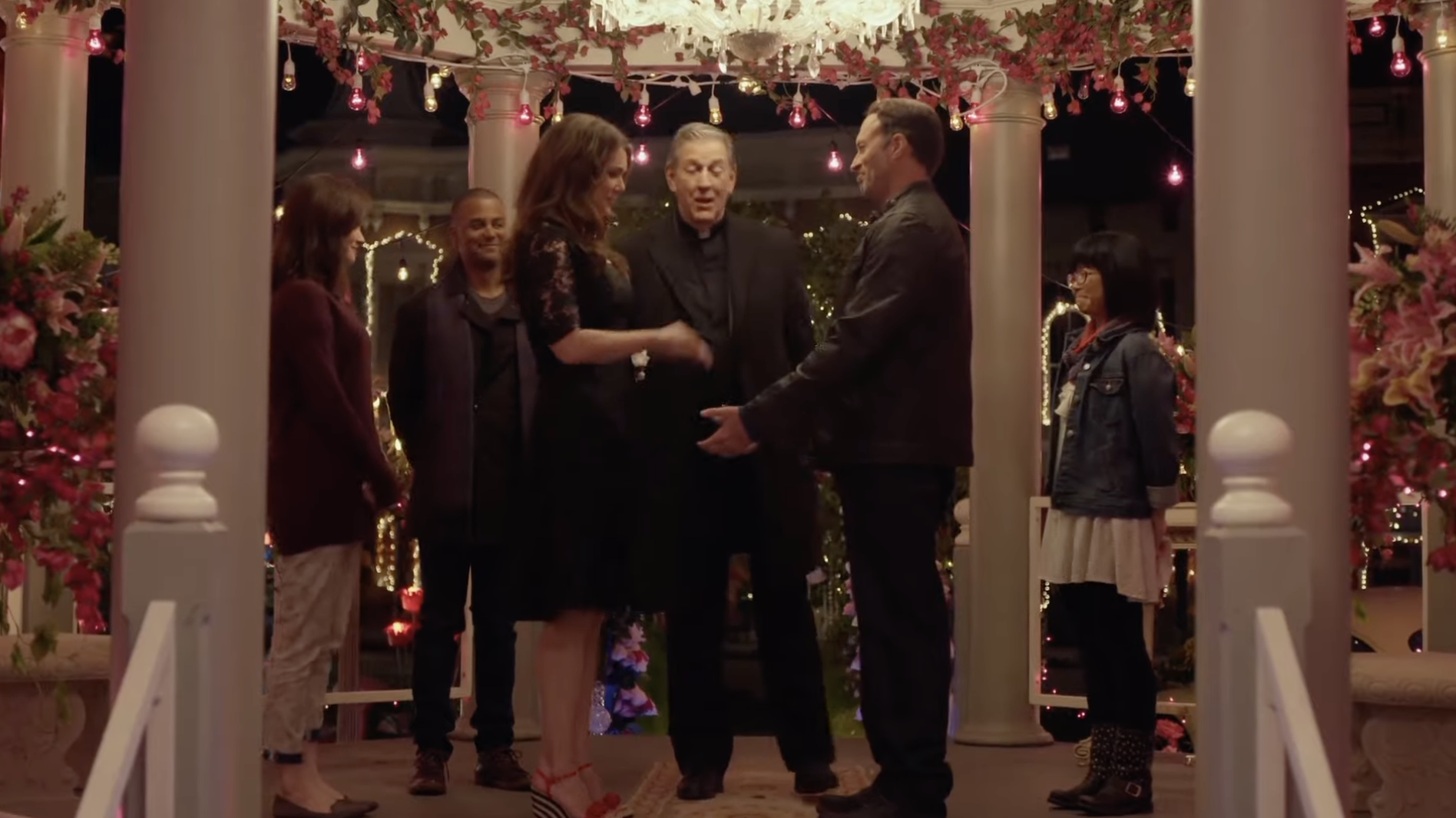 Lorelai and Luke stand hand in hand in the center of a gazebo which has been decorated with flowers and string lights