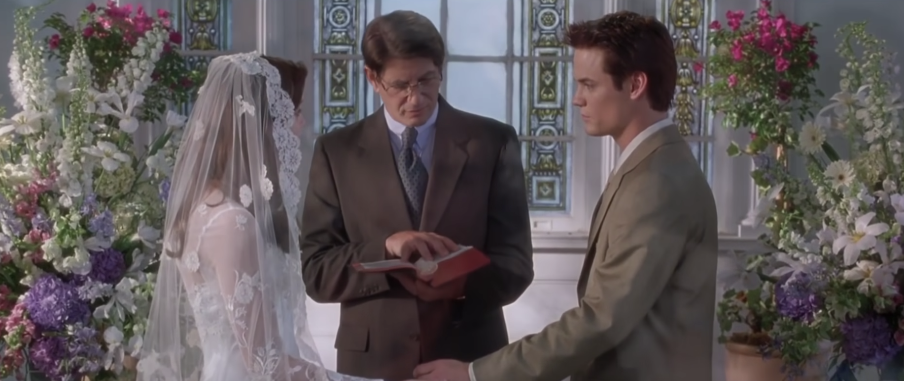 A bride and groom stand at the altar, she has a traditional veil covering her face and he is in a khaki suit - they&#x27;re surrounded by flowers and an officiant stands between them reading from a book