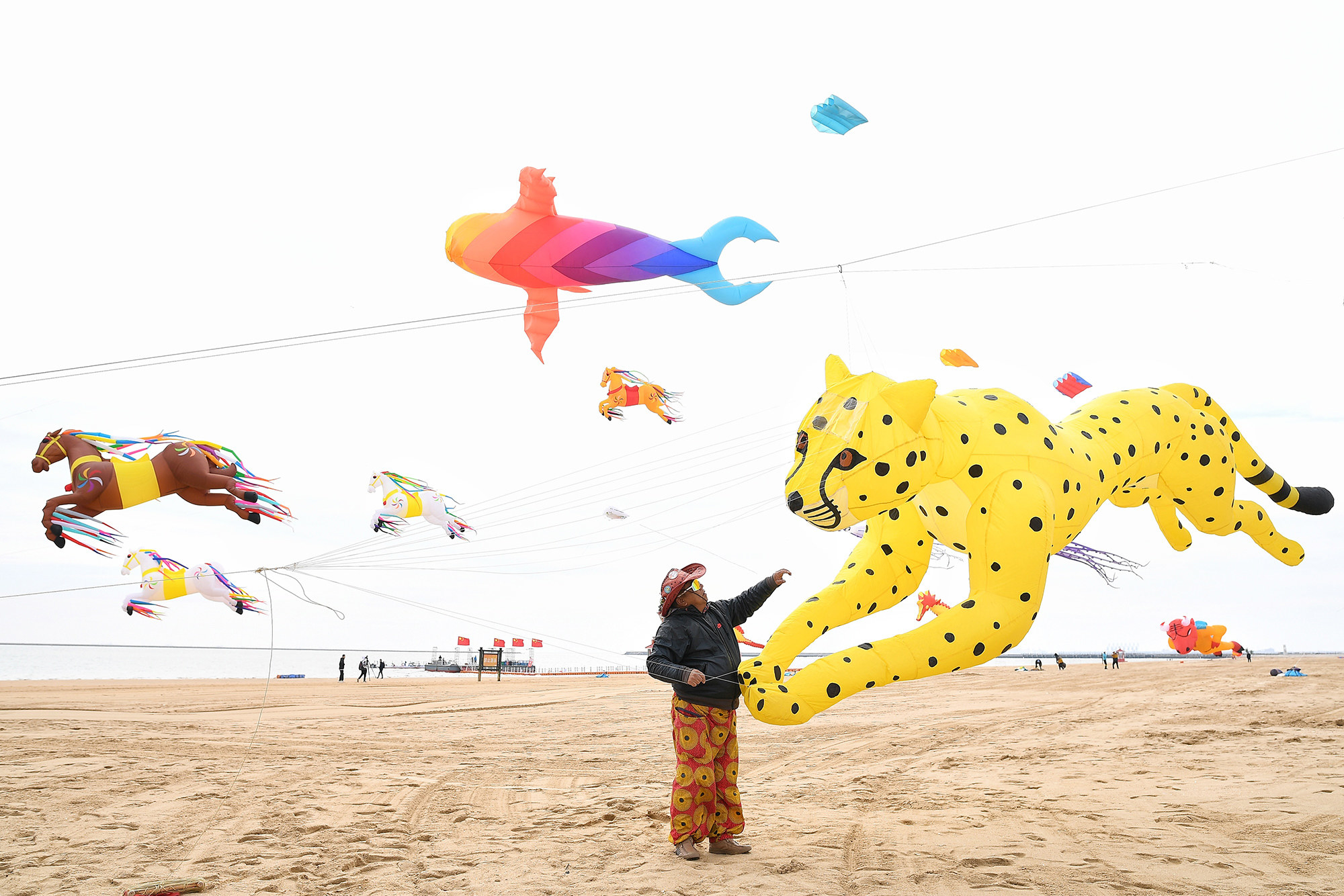 someone flies a gigantic yellow leopard kite in an openfield; in the background, there are colorful fish and horse kites in the air