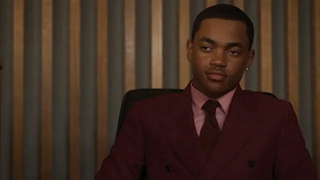 Michael Rainey Jr as Tariq sitting and wearing a colorful suit and tie and staring