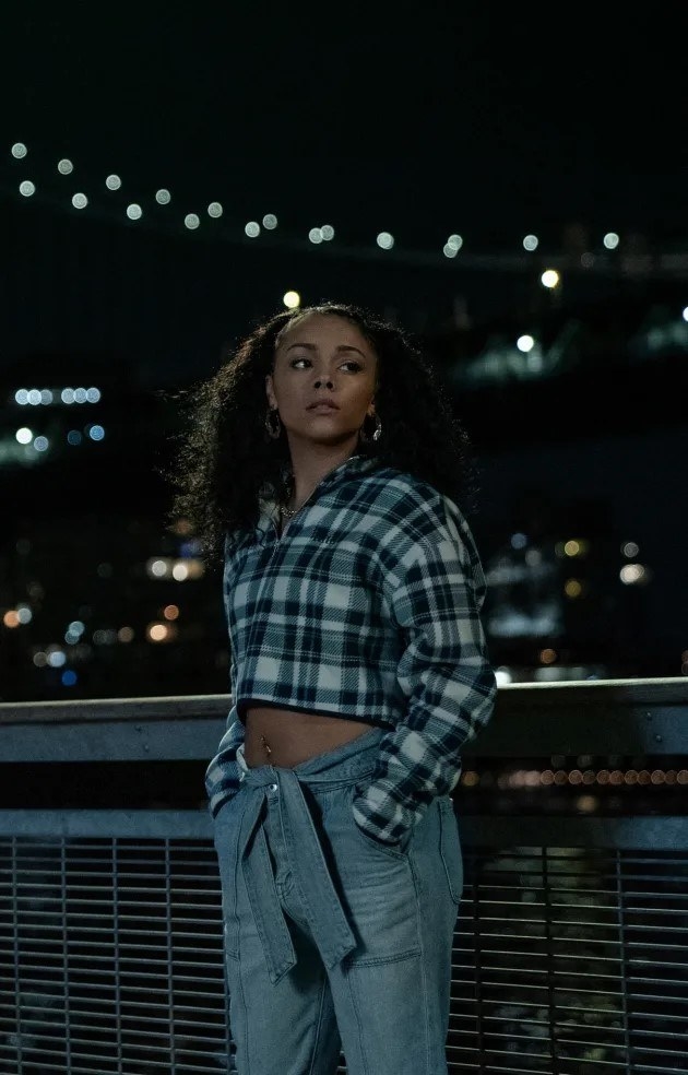 Effie Waits in a cutoff flannel top and jeans, standing in front of a bridge
