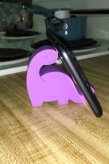 Reviewer's tiny purple dino shaped phone stand holding up a phone on a counter