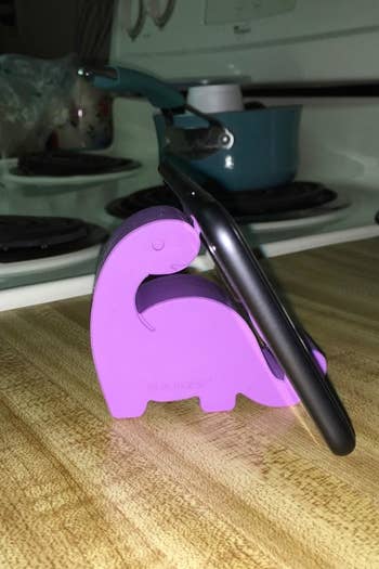 Reviewer's tiny purple dino shaped phone stand holding up a phone on a counter