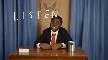 GIF of a young man, sitting behind a wooden desk, saying &quot;listen listen listen&quot;.