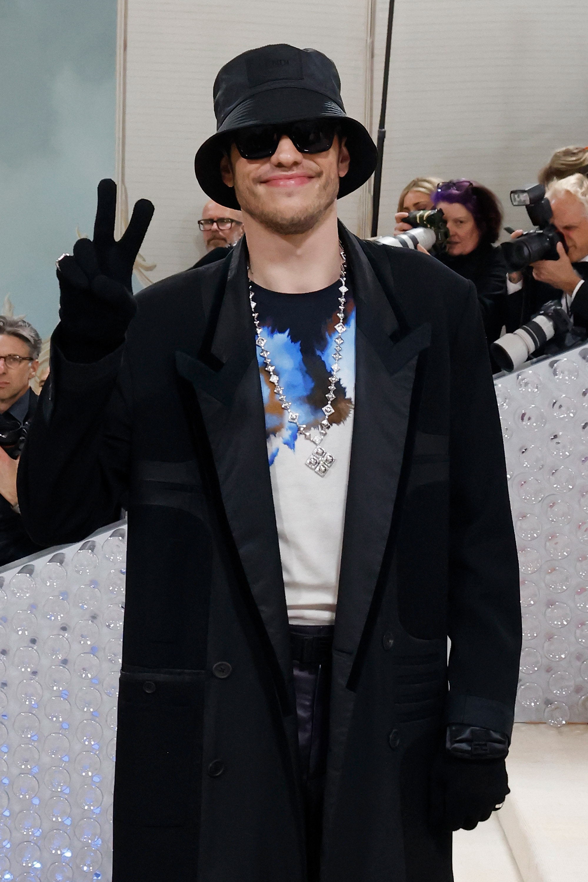Pete giving the peace sign as he stands at a red carpet event. He&#x27;s wearing a long coat, gloves, bucket hat, sunglasses, and a chain