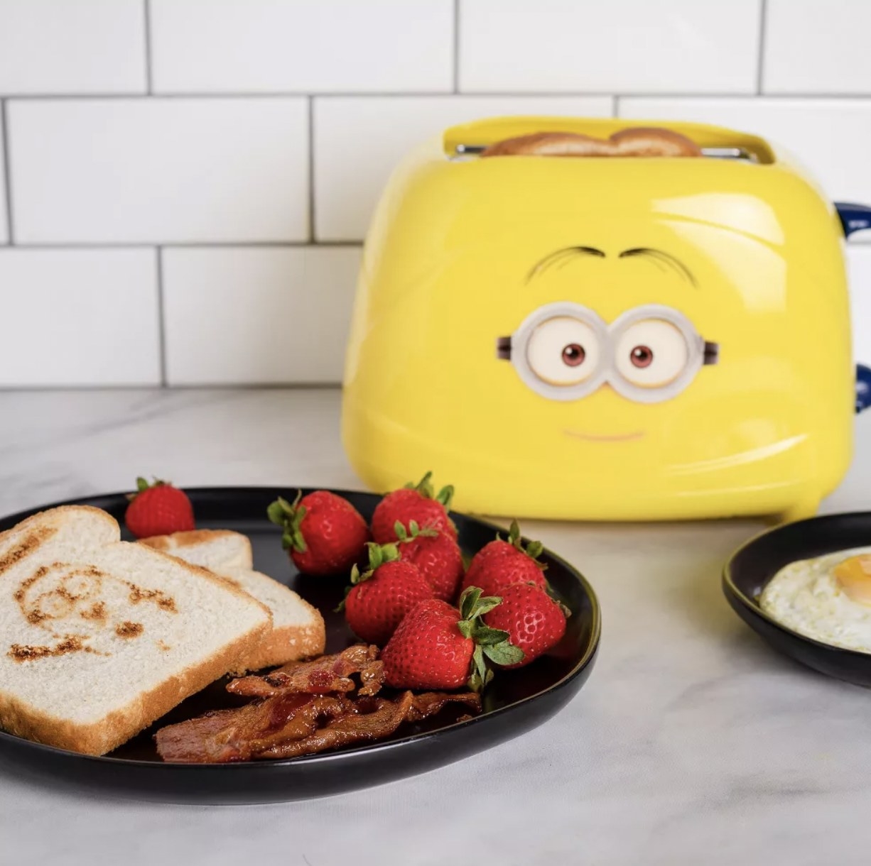 the toaster next to plate with minion-printed white toast, bacon, and strawberries