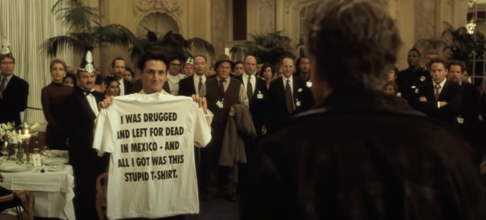A man at a party holding up a shirt that reads: &quot;I was drugged and left for dead in Mexico, and all I got was this stupid t-shirt&quot;