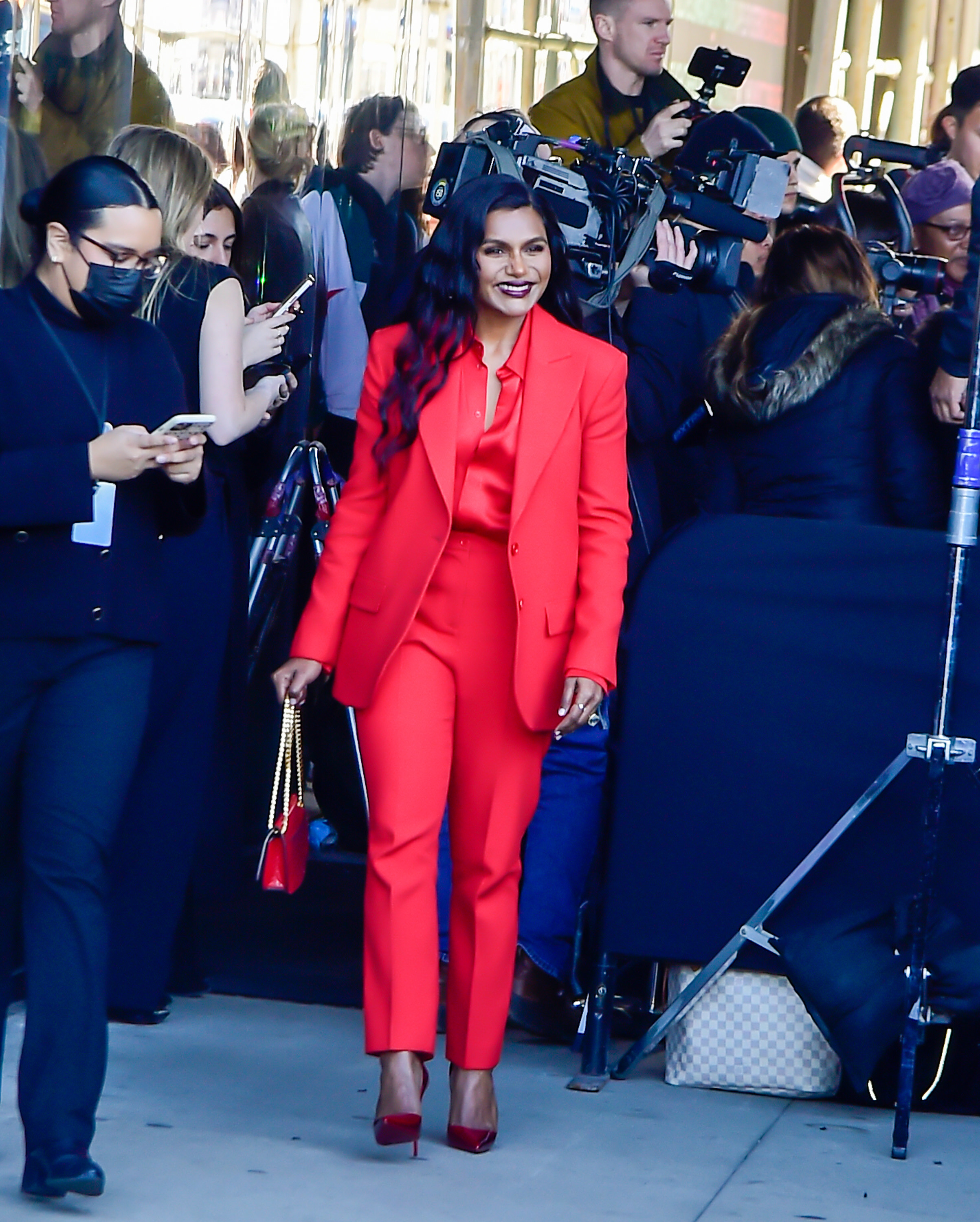 Mindy walks out of a building in a pantsuit with matching bag and stilletto heels