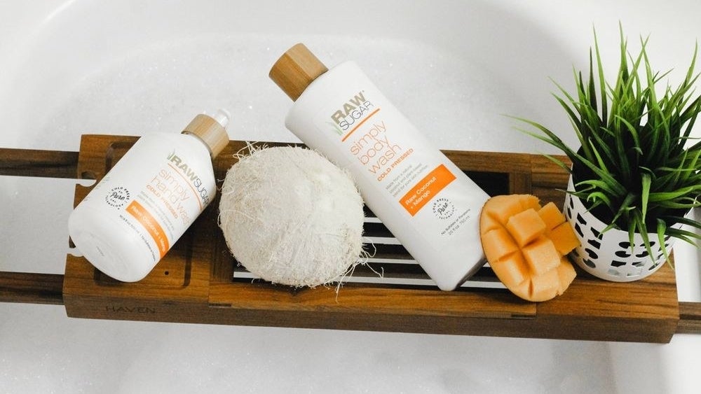 A picture of the body wash product beside another product from the line, a coconut and a mango in a bathtub