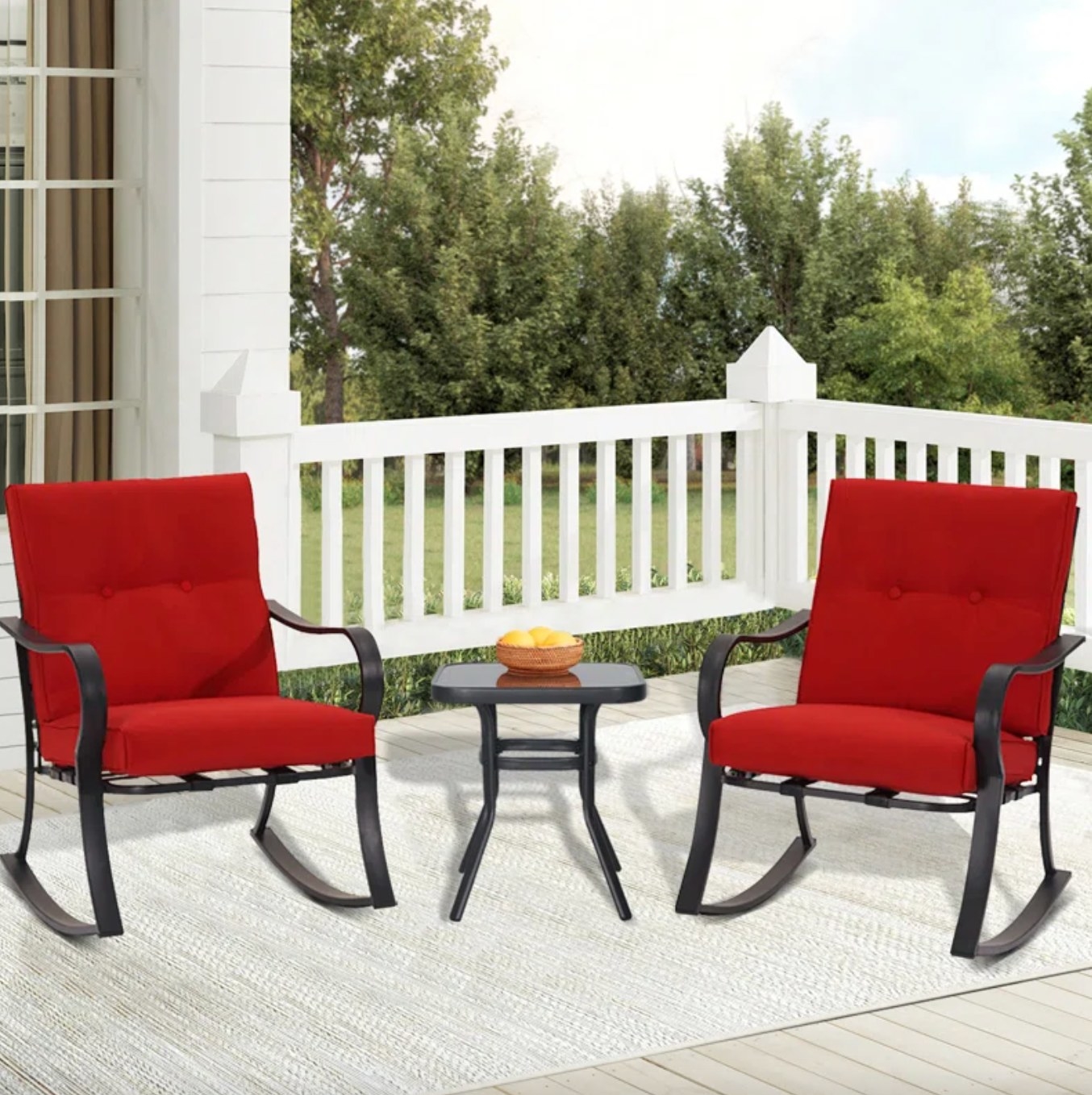 the black metal rocking chairs and small table. the chairs have red cushions, and the whole set is on a white porch in front of a yard and wooded area