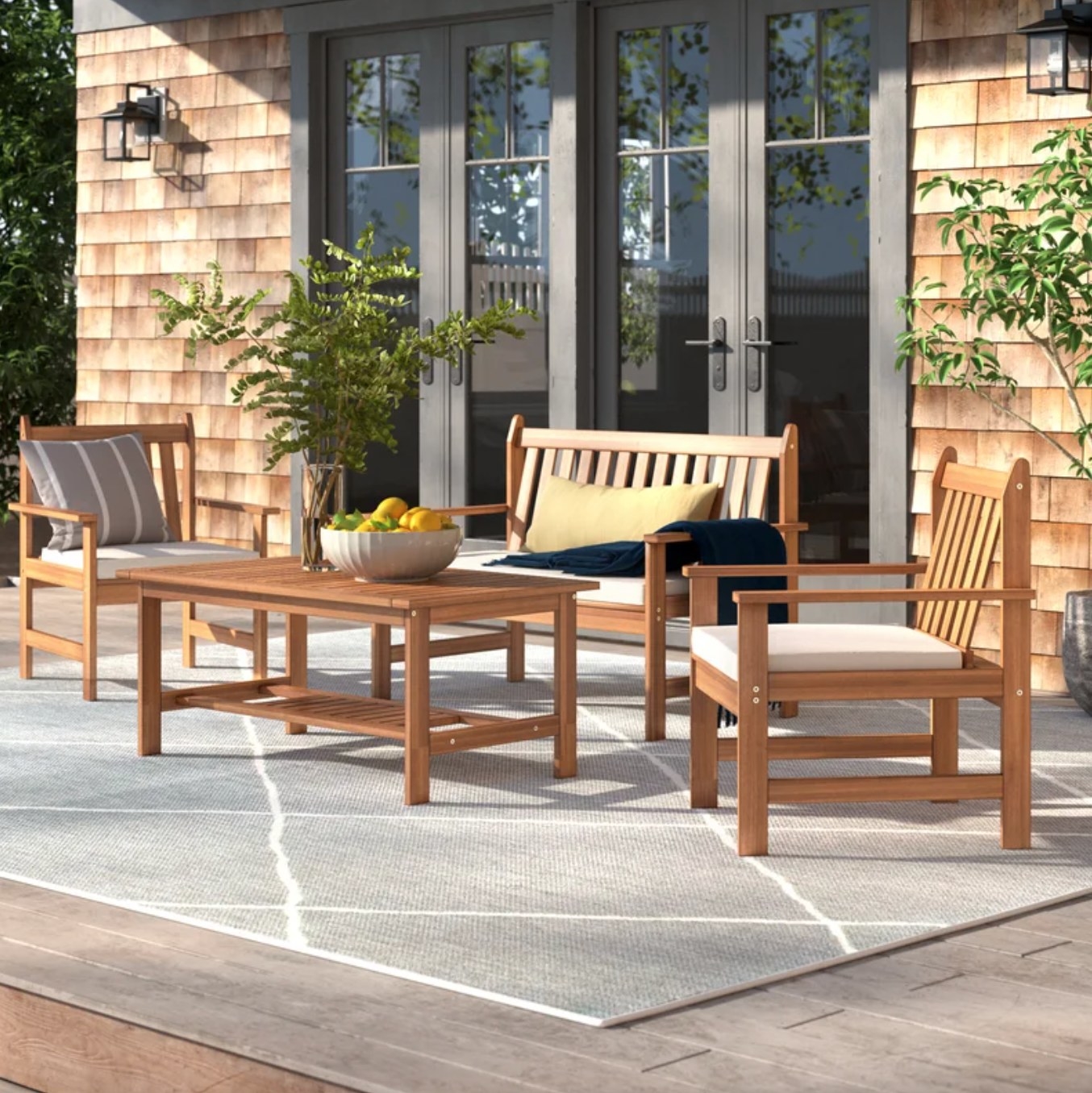 the light wood coffee table, chairs and loveseat. the chairs and sofa have light cushions, and the whole set is on a light outdoor rug on a deck