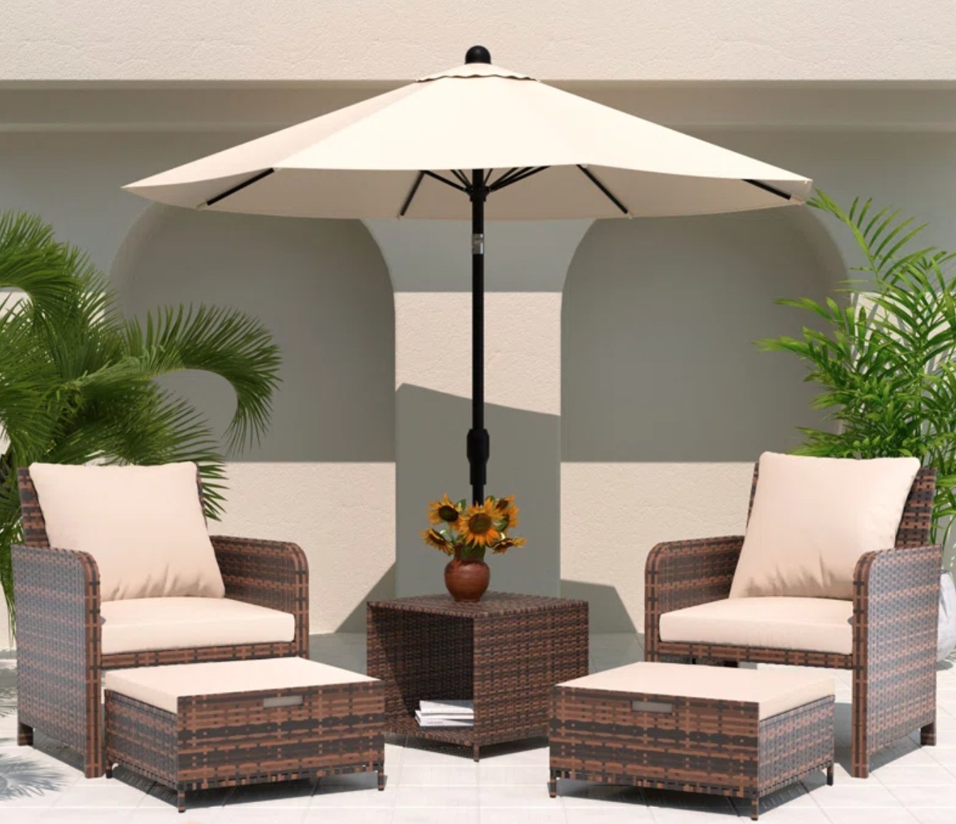 the dark wicker chairs, ottomans and side table with beige cushions on the chairs and ottomans. The set is on a patio with a large umbrella