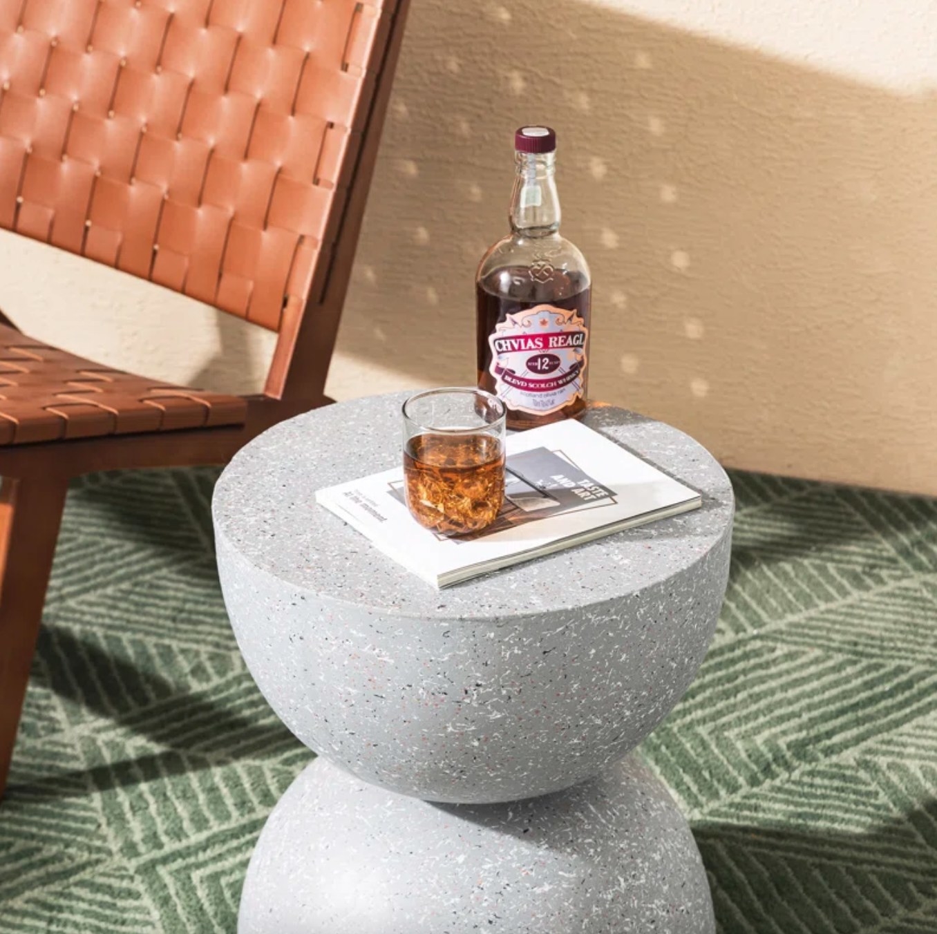 the grey hourglass shaped stool next to an outdoor chair with a drink and a book on top