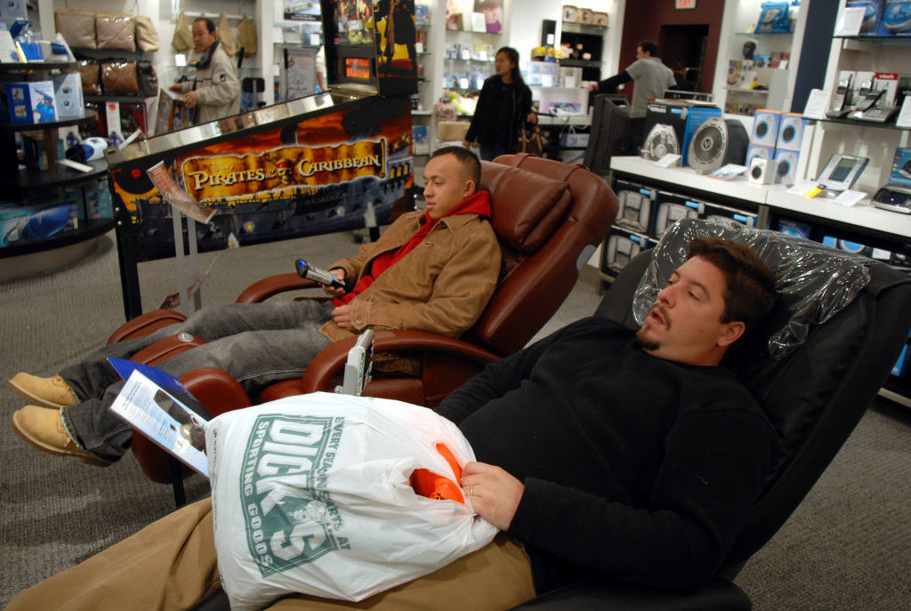 Two men sitting in the massage chairs with a pinball machine right next to them