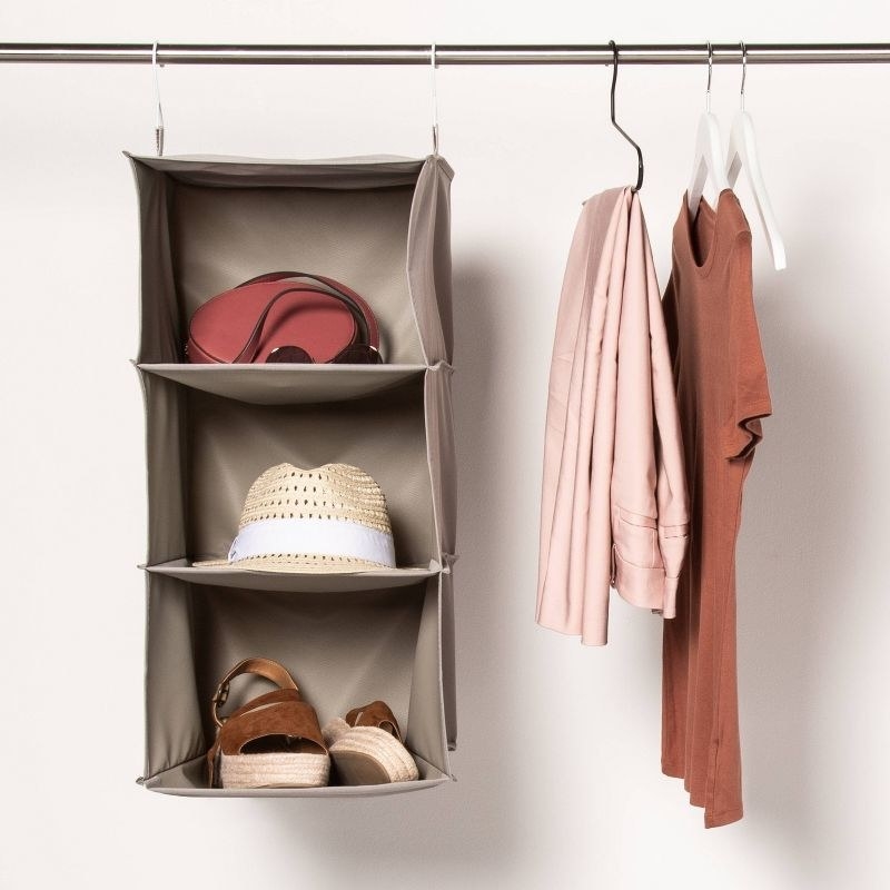 a gray hanging closest organizer with three shelves holding a hat, shoes, and purse