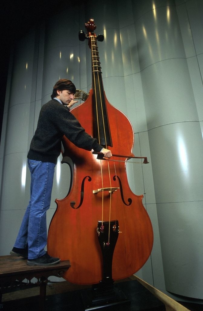 A man standing on a step alongside a huge, cello-like instrument and playing it, and it&#x27;s still taller than he is