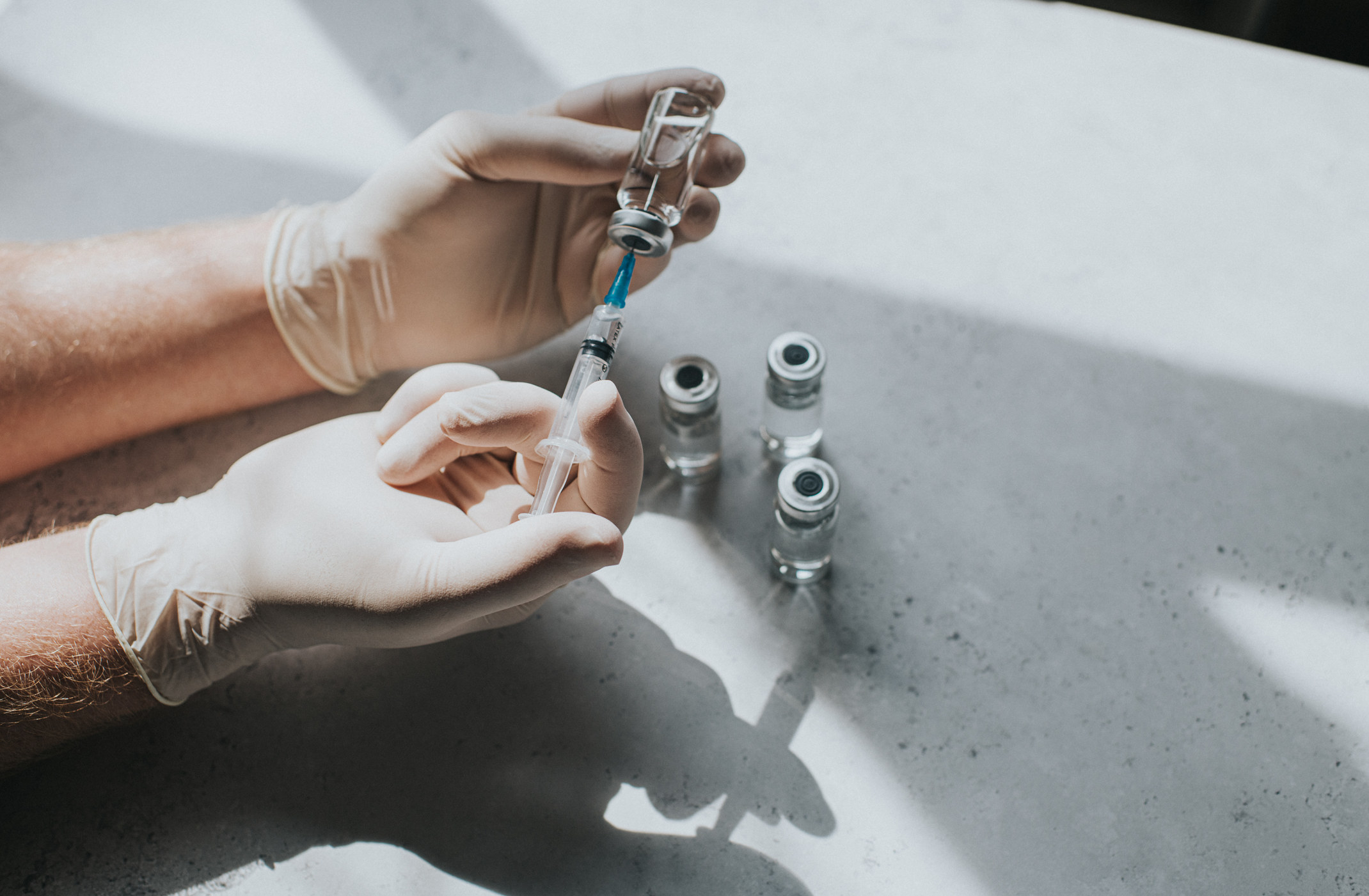 A hand wearing latex gloves hold a phial of clear liquid and a syringe