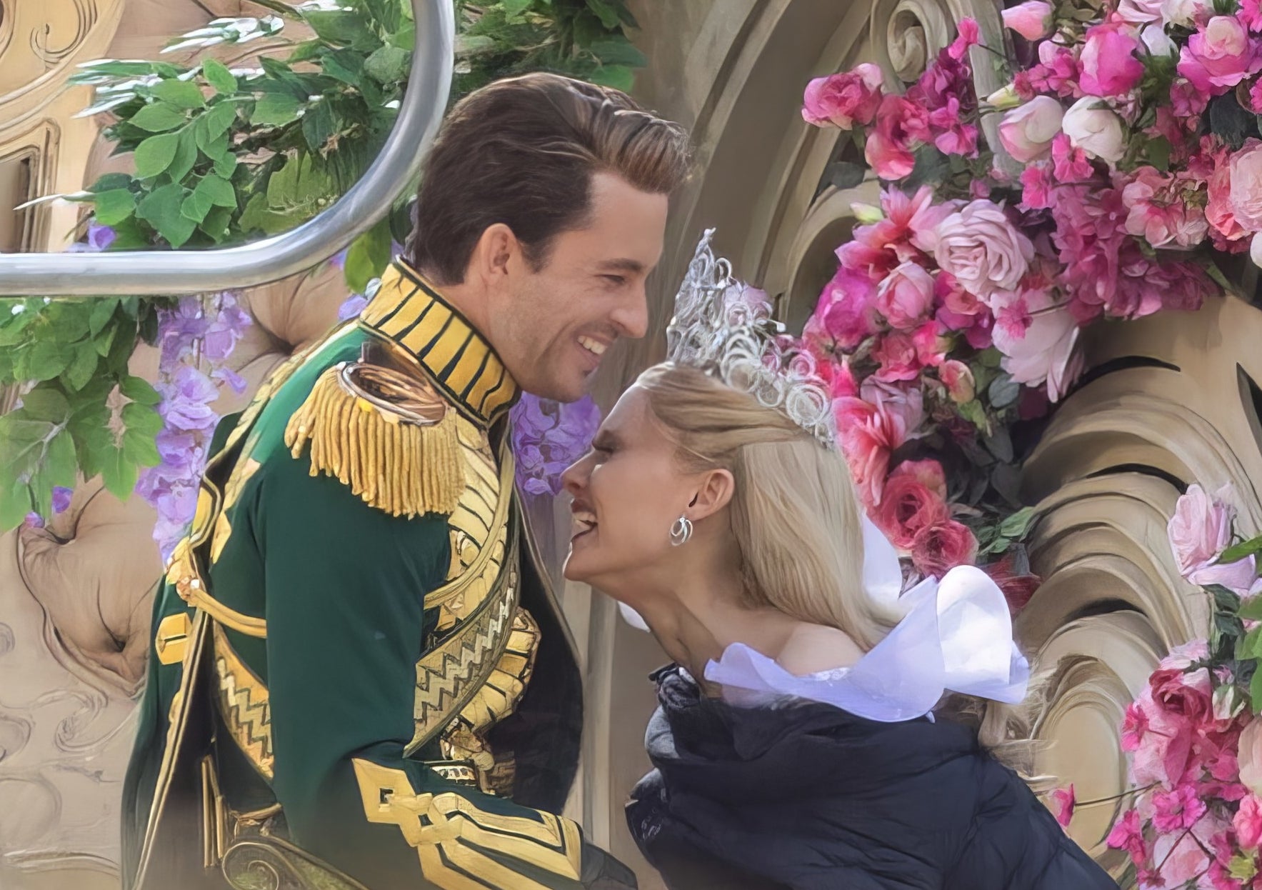 jonathan bailey and ariana grande laugh on the set of wicked in front of a set that is decorated with with foliage and flowers
