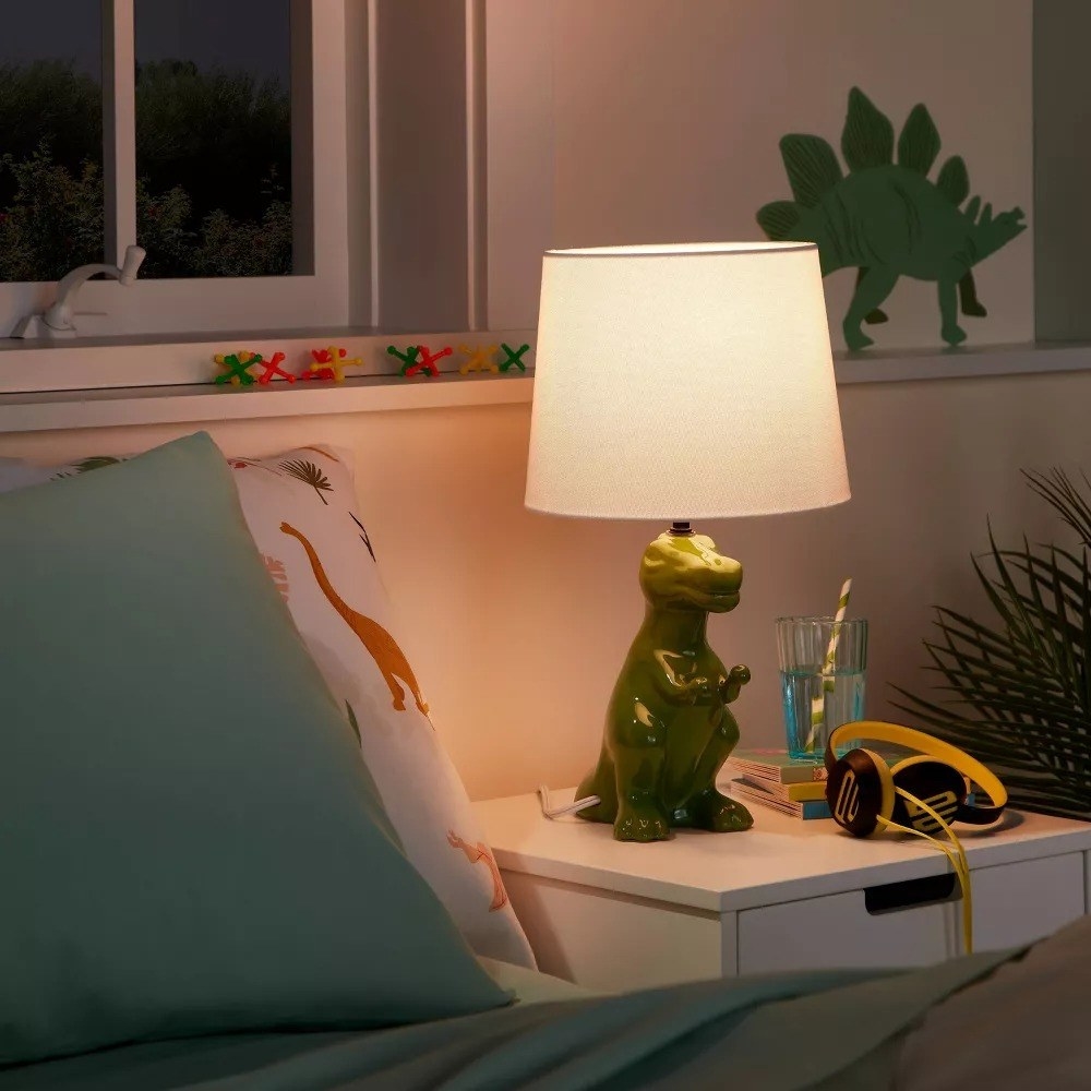 lit green T-Rex lamp on a nightstand next to a bed