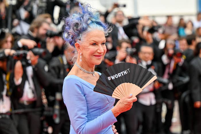 Helen has her purple hair in pinned curls on top of her head while holding a paper fan