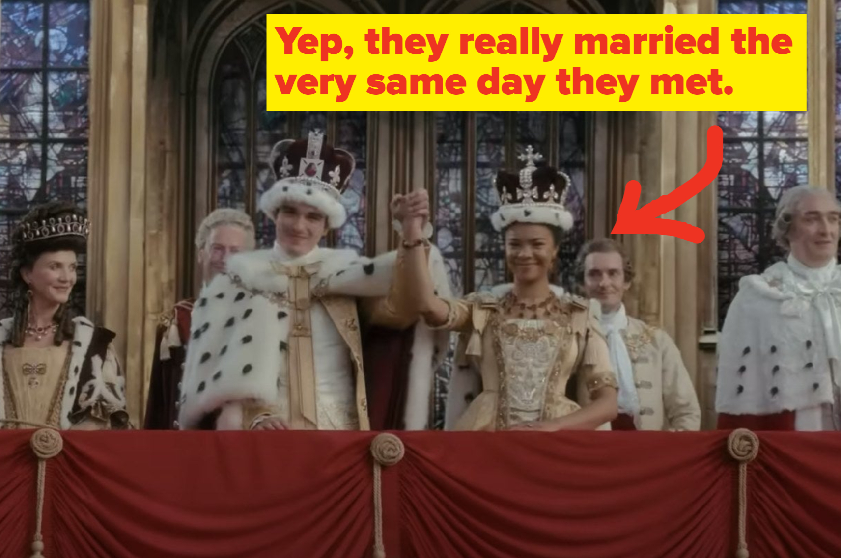 In a scene from 'Queen Charlotte: A Bridgerton Story" King George III and Queen Charlotte hold their raised hands on a castle balcony surrounded by dignitaries