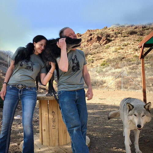 A man and a woman in jeans outdoors with two large dogs, one resting on their shoulders