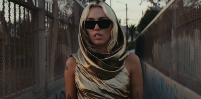 A closeup of Miley Cyrus walking in a metallic outfit and sunglasses in her video for Flowers
