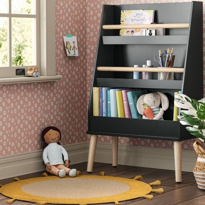 a charcoal grey three-shelf bookshelf filled with books in a kid&#x27;s room with a sun rug and a plush doll