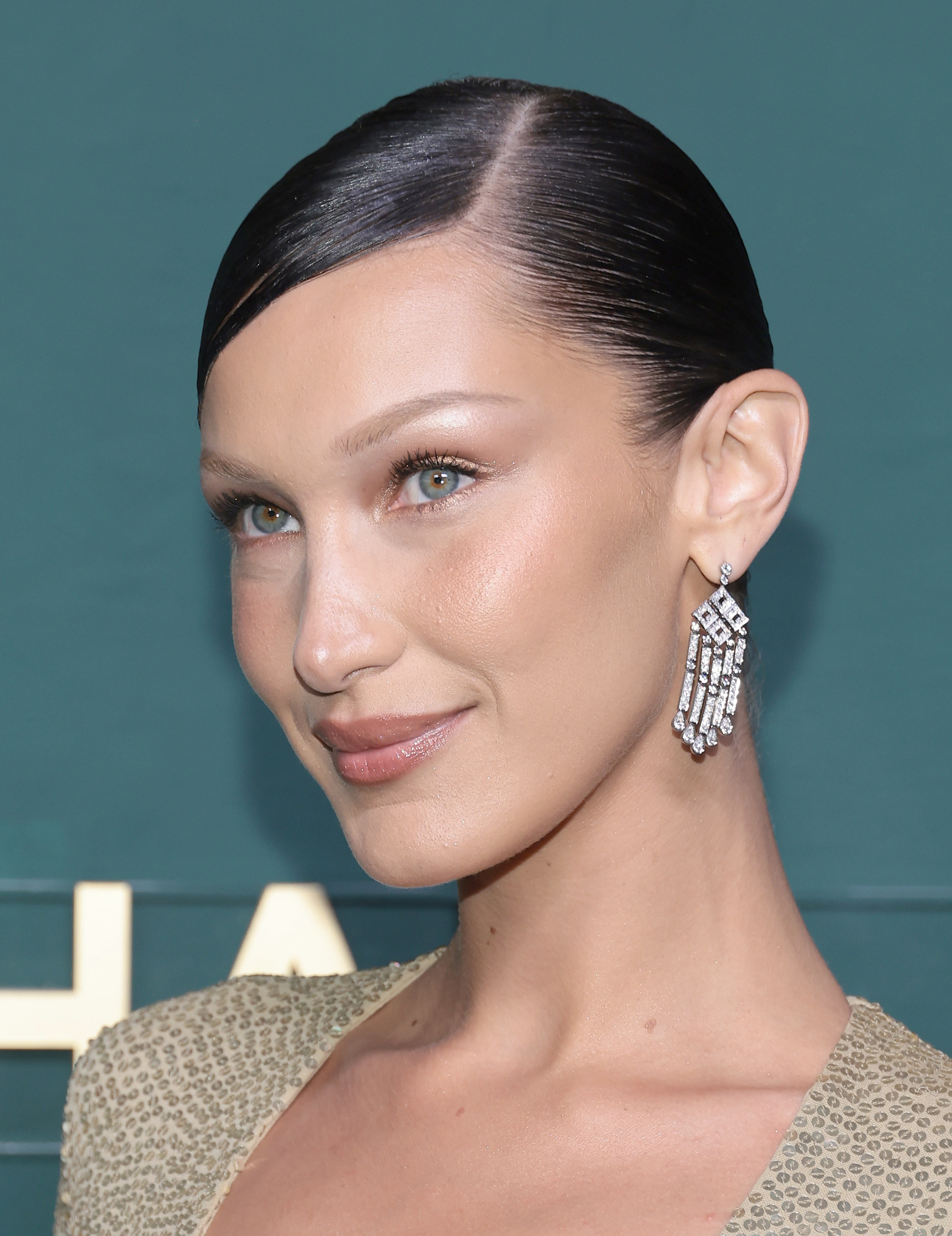 A close-up photo of Bella Hadid smiling with a slick bun, sequined shirt, and big diamond earrings